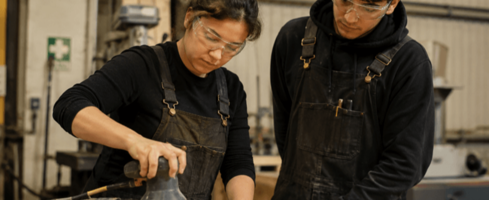 A woman with safety glasses uses a hand sanding tool on a piece of wood while man watches closely beside her.